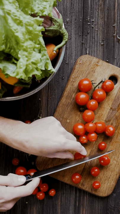 Hands slicing cherry tomatoes on a board beside a bowl of salad